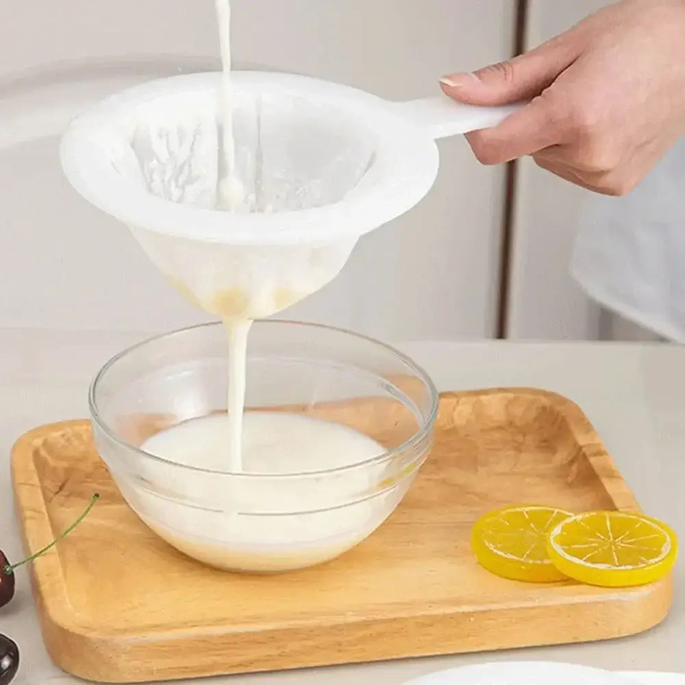 Hand pouring liquid through ultra fine nylon filter mesh strainer into a glass bowl on a wooden tray with lemon slices.