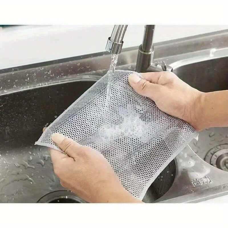 Person washing a 12pcs non-stick oil iron dishwashing cloth under running water in a kitchen sink.