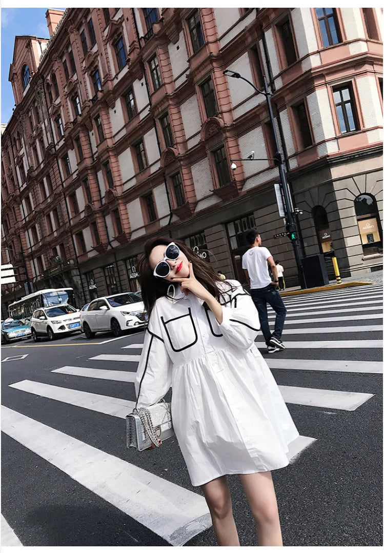 Woman in a stylish white shirt dress with pockets, crossing the street in a chic urban setting.