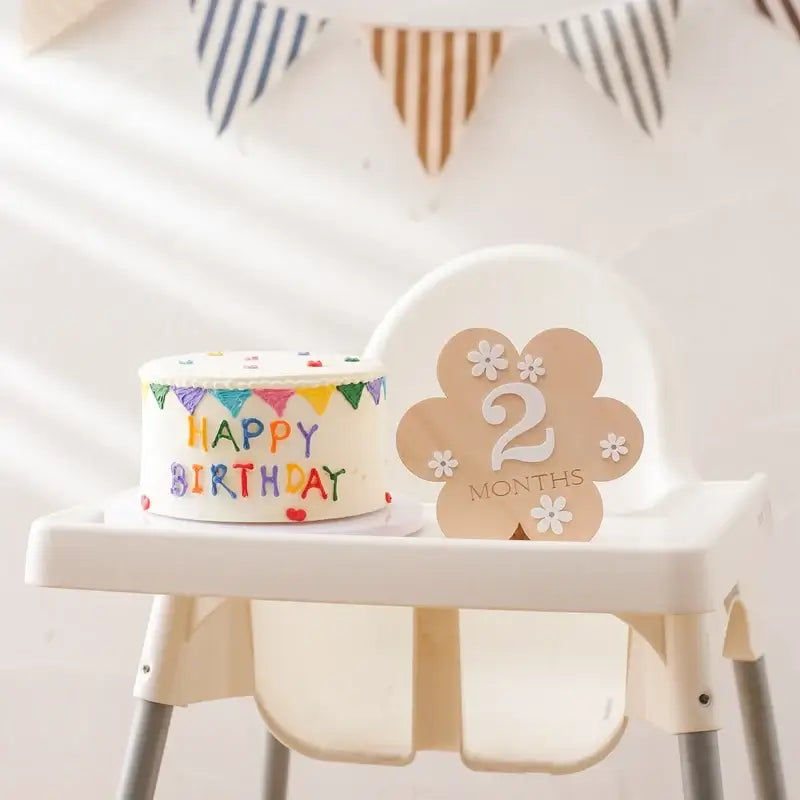 Wooden baby milestone flower shape displaying '2 Months' next to a colorful birthday cake on a high chair.
