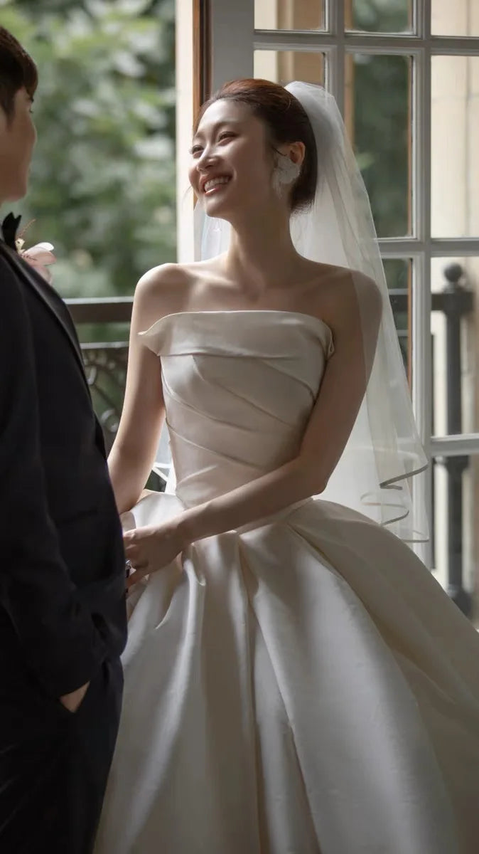 Bride wearing a satin off-shoulder wedding dress, smiling with a veil in a romantic setting.