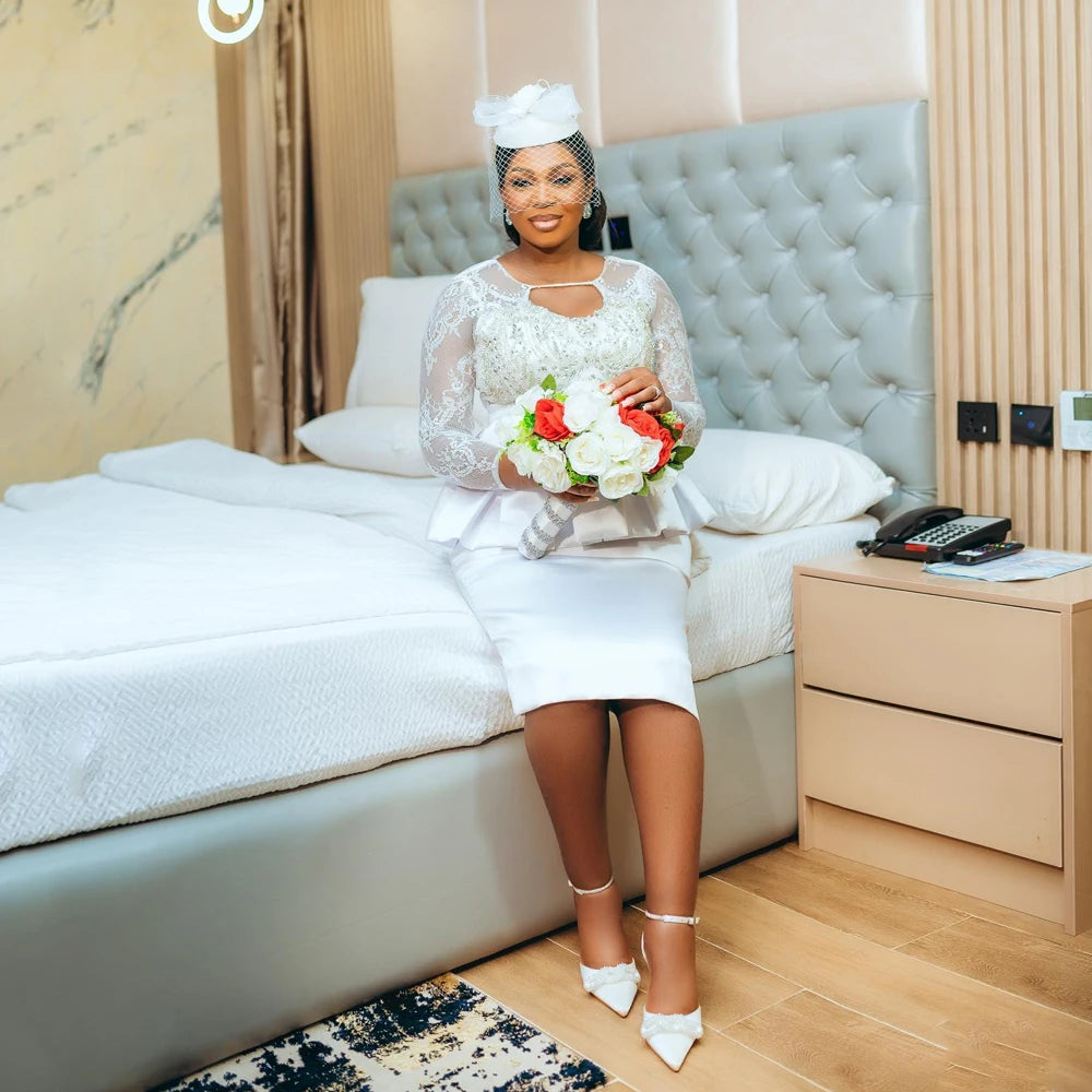 Elegant bride in a lace and satin wedding dress, holding a bouquet, sitting by a modern bed in a stylish room.