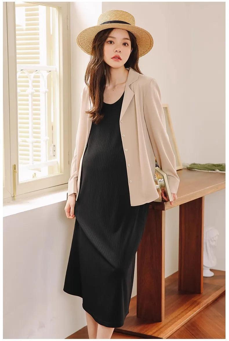 Maternity model wearing a sleeveless black dress with a beige blazer and straw hat in a bright, airy room.