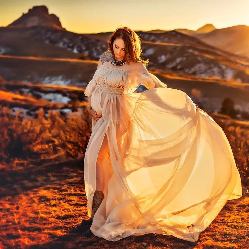 Expectant mother in a flowing boho maternity dress against a sunset backdrop in a natural landscape.