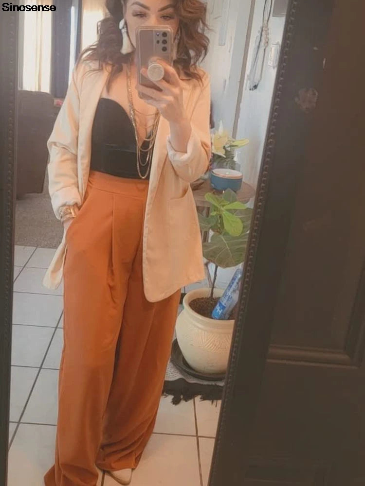 Woman posing in mirror wearing a black bodysuit, beige blazer, and orange wide-leg pants.
