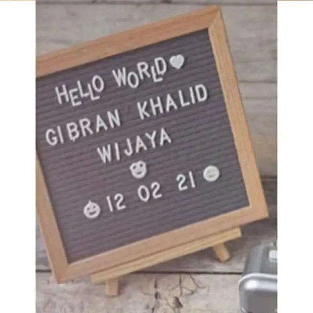Letter board displaying "Hello World Gibran Khalid Wijaya 12 02 21" with decorative emojis on a wooden background.