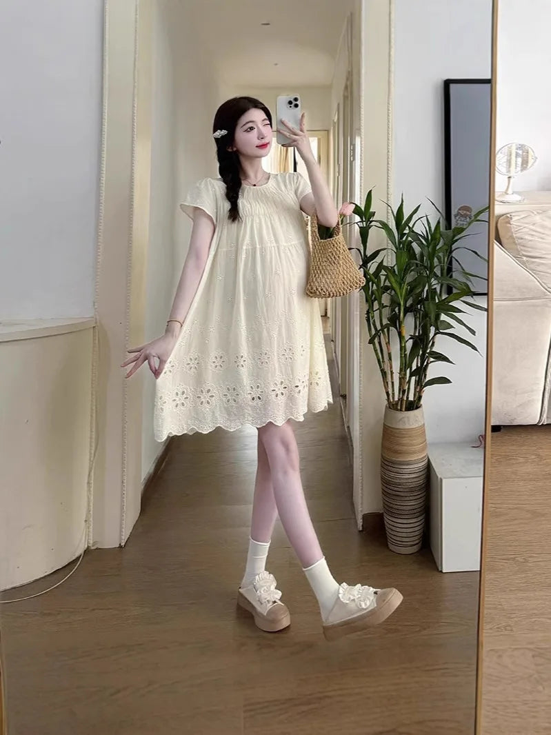 Model showcasing a knee-length A-line cotton maternity dress in cream, styled with a woven bag, set in a modern interior.