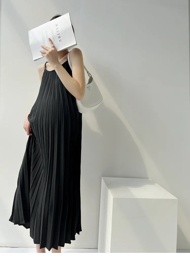 Model wearing a pleated black maternity dress, holding a book, against a minimalist background.