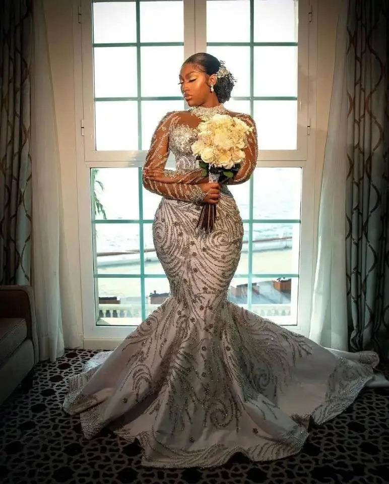 Luxury mermaid wedding dress with beaded sheer sleeves, model holding bouquet near window, elegant bridal style.