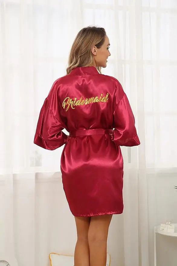 Back view of a bridesmaid wearing a satin robe with golden lettering in a bright, airy room.