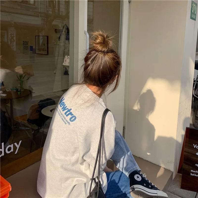 Woman wearing oversized gray Harajuku streetwear tee and jeans, sitting at a cafe.