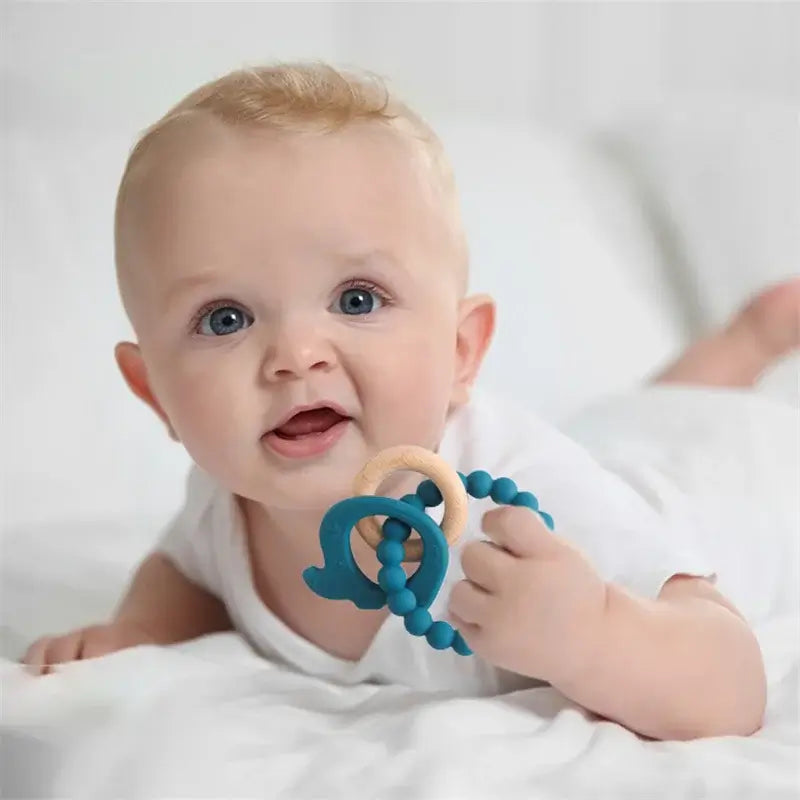 Smiling baby lying on a bed, holding a blue silicone teether and wooden ring, showcasing safe and non-toxic materials.