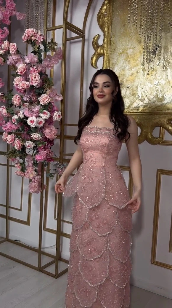 Elegant woman in a pink beaded engagement gown with a tiered skirt, standing near pink flower decor.