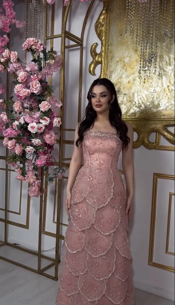 Customized pink beaded engagement dress with tiered skirt and lace-up back, featuring a floral backdrop.