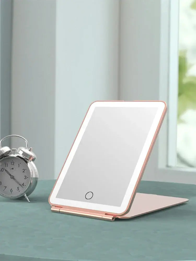 LED foldable travel makeup mirror with USB rechargeable feature on a table beside an alarm clock.