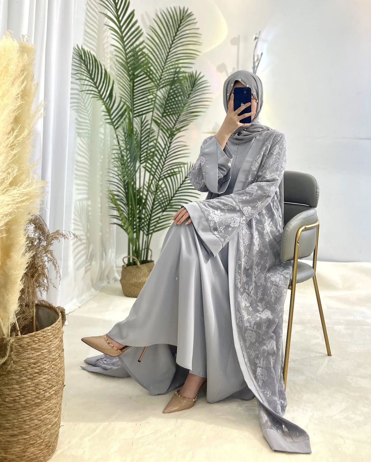 Model wearing a stylish Dubai Moroccan Kaftan dress in gray, seated in a modern setting with greenery.