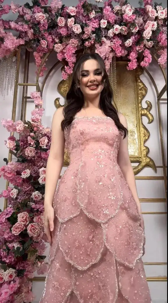 Customized pink beaded engagement dress with tiered skirt, featuring a lace-up back and floral backdrop.