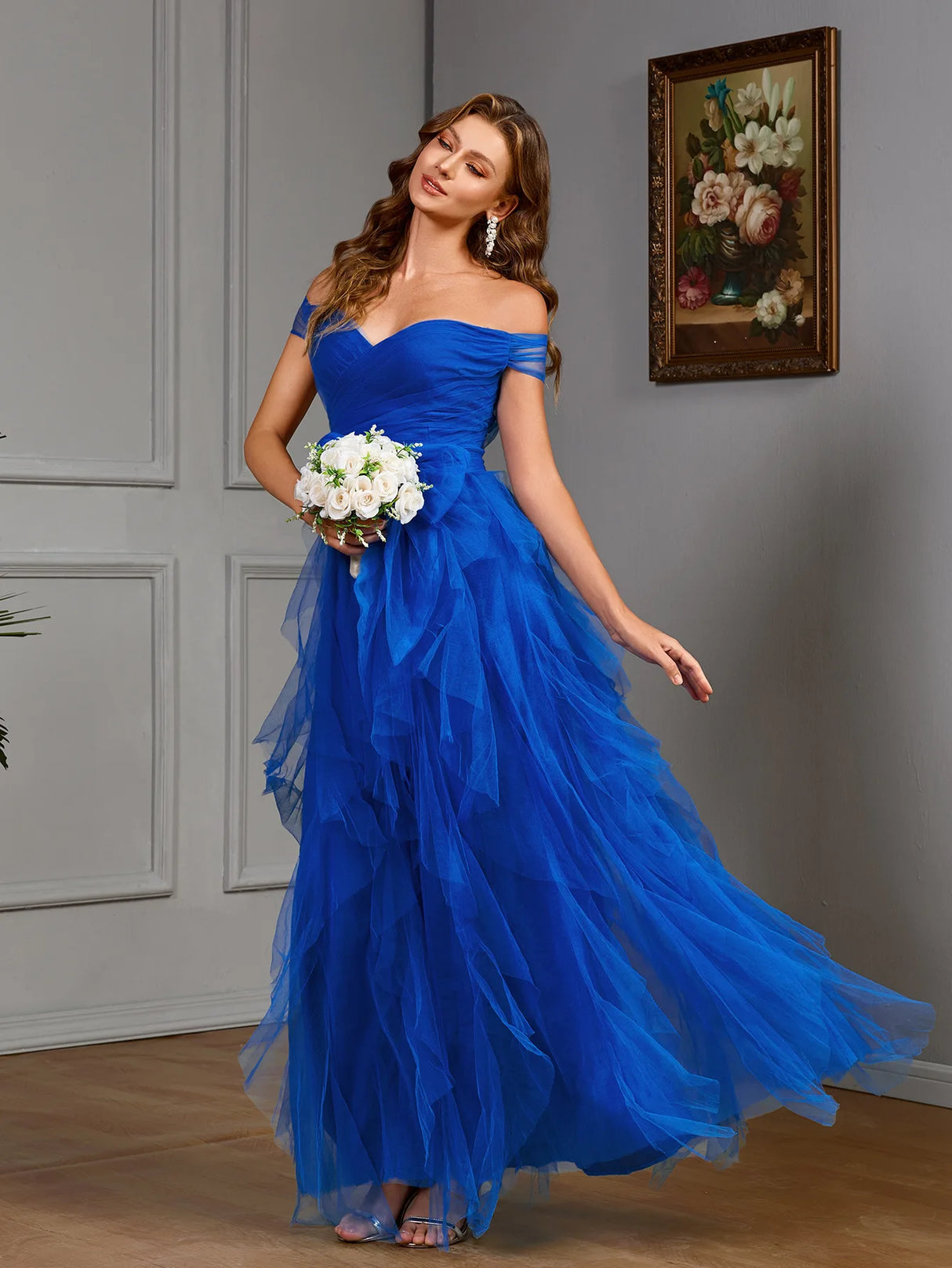 Model wearing a vibrant blue off-the-shoulder gown with layered tulle, holding a bouquet in an elegant setting.