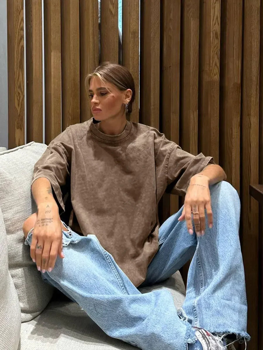 Woman sitting on a couch wearing a brown oversized t-shirt and light blue jeans, relaxed in a modern setting.