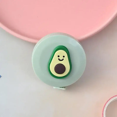 Cute avocado-shaped tape measure with a smiling face on a light green casing, placed on a pink background.
