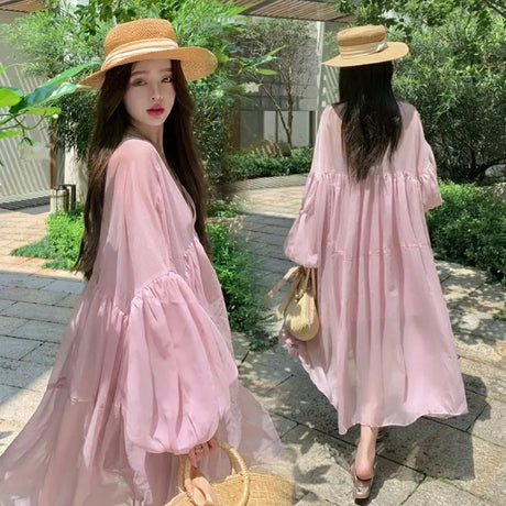 Woman in a pink chiffon strappy maternity dress with bell sleeves, wearing a straw hat in a sunny outdoor setting.