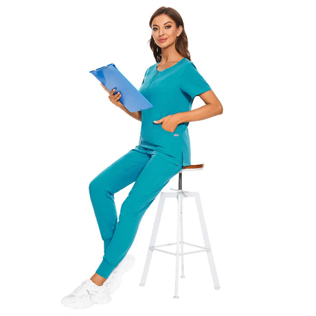 Woman in turquoise nursing work uniform with zipper V-neck top and pockets, sitting on stool with clipboard.