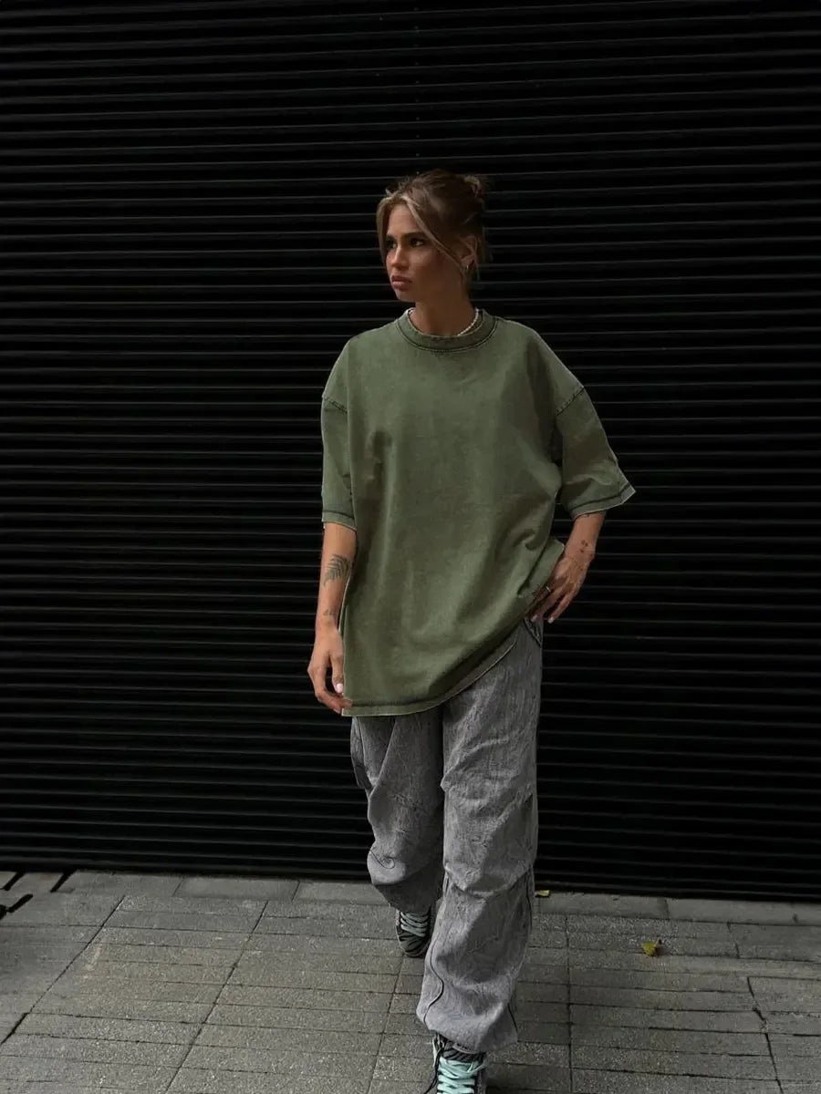 Woman wearing an oversized green t-shirt and loose grey pants, walking against a black slatted background.