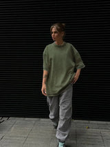 Woman wearing an oversized green t-shirt and loose grey pants, walking against a black slatted background.