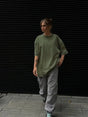Woman wearing an oversized green t-shirt and loose grey pants, walking against a black slatted background.