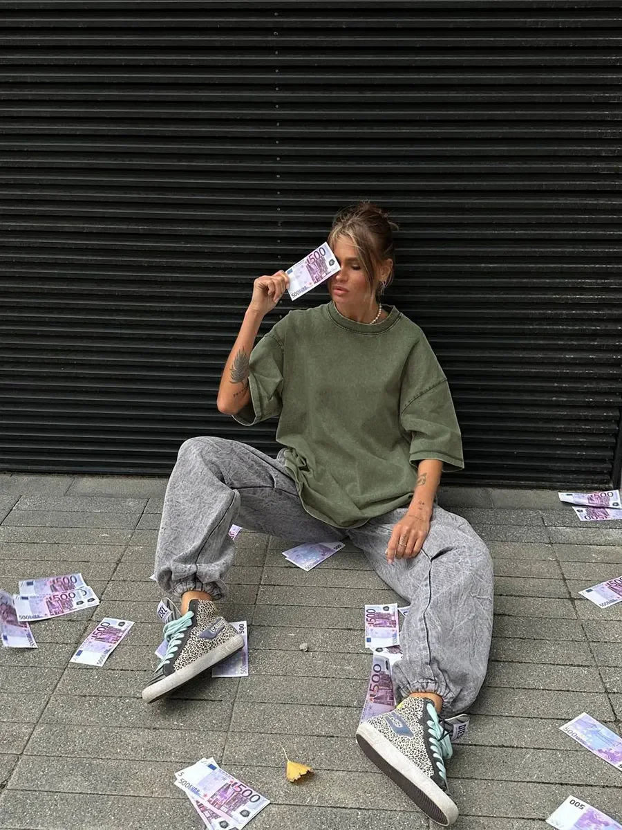 Woman wearing a green oversized t-shirt and gray pants, sitting on the ground with fake money around her.