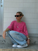 Woman wearing a pink casual t-shirt and baggy jeans, sitting stylishly outdoors with sunglasses.