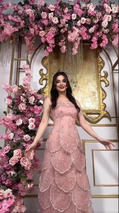 Pink beaded engagement gown with tiered skirt, lace-up back, and floral backdrop. Perfect for weddings and special occasions.