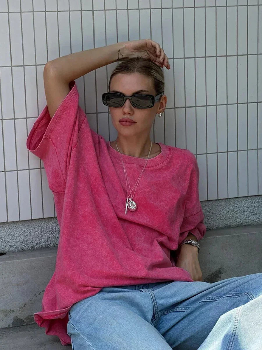 Model wearing a pink Wolfeel wax printed casual t-shirt, sitting against a tiled wall in relaxed jeans and sunglasses.
