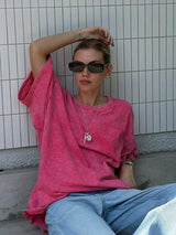 Model wearing a pink Wolfeel wax printed casual t-shirt, sitting against a tiled wall in relaxed jeans and sunglasses.