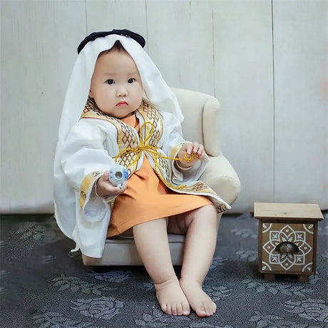 Baby boy in a creative mini suitcase outfit, wearing a white robe with gold accents and sitting adorably on a small chair.